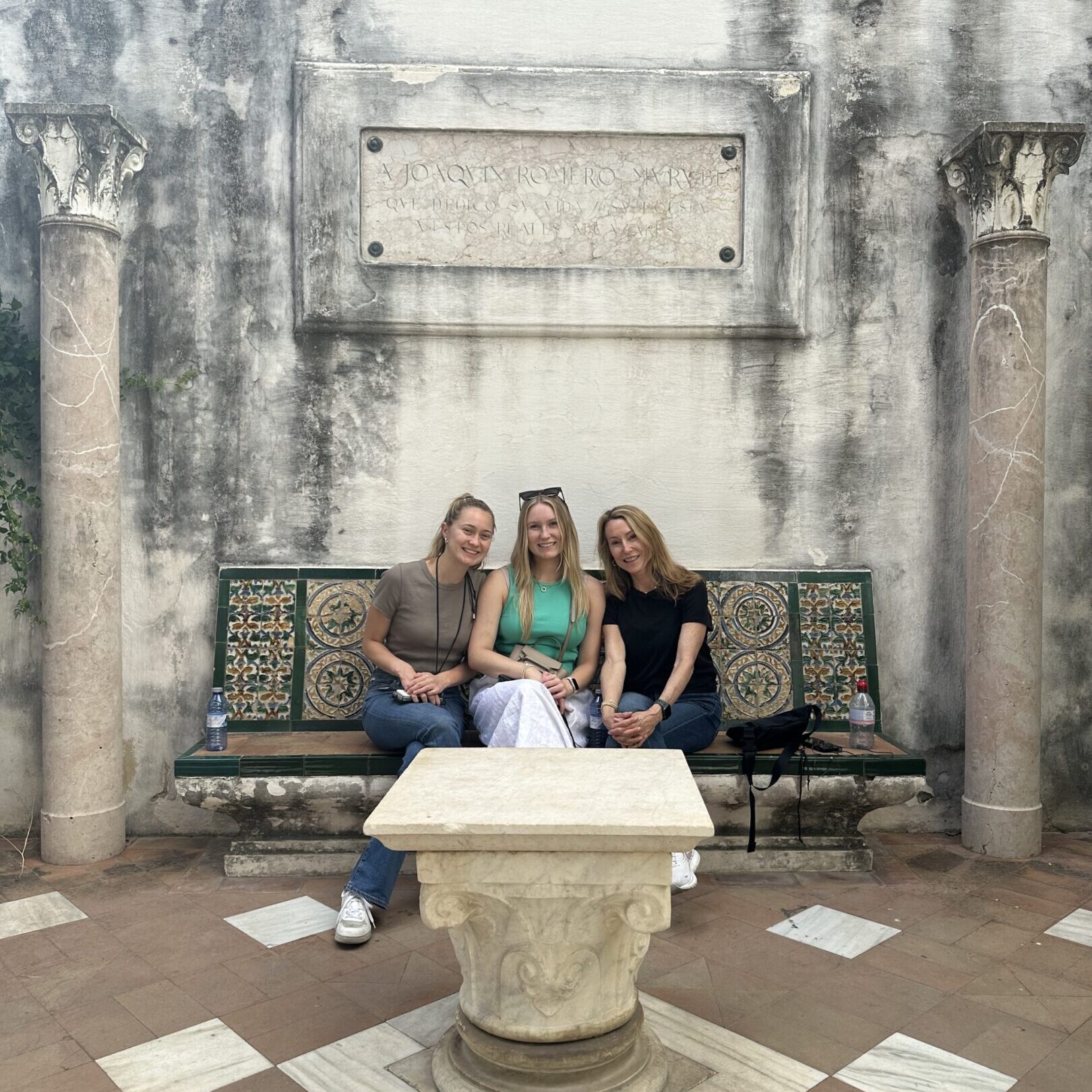 Three women sitting on a bench in an old stone courtyard.